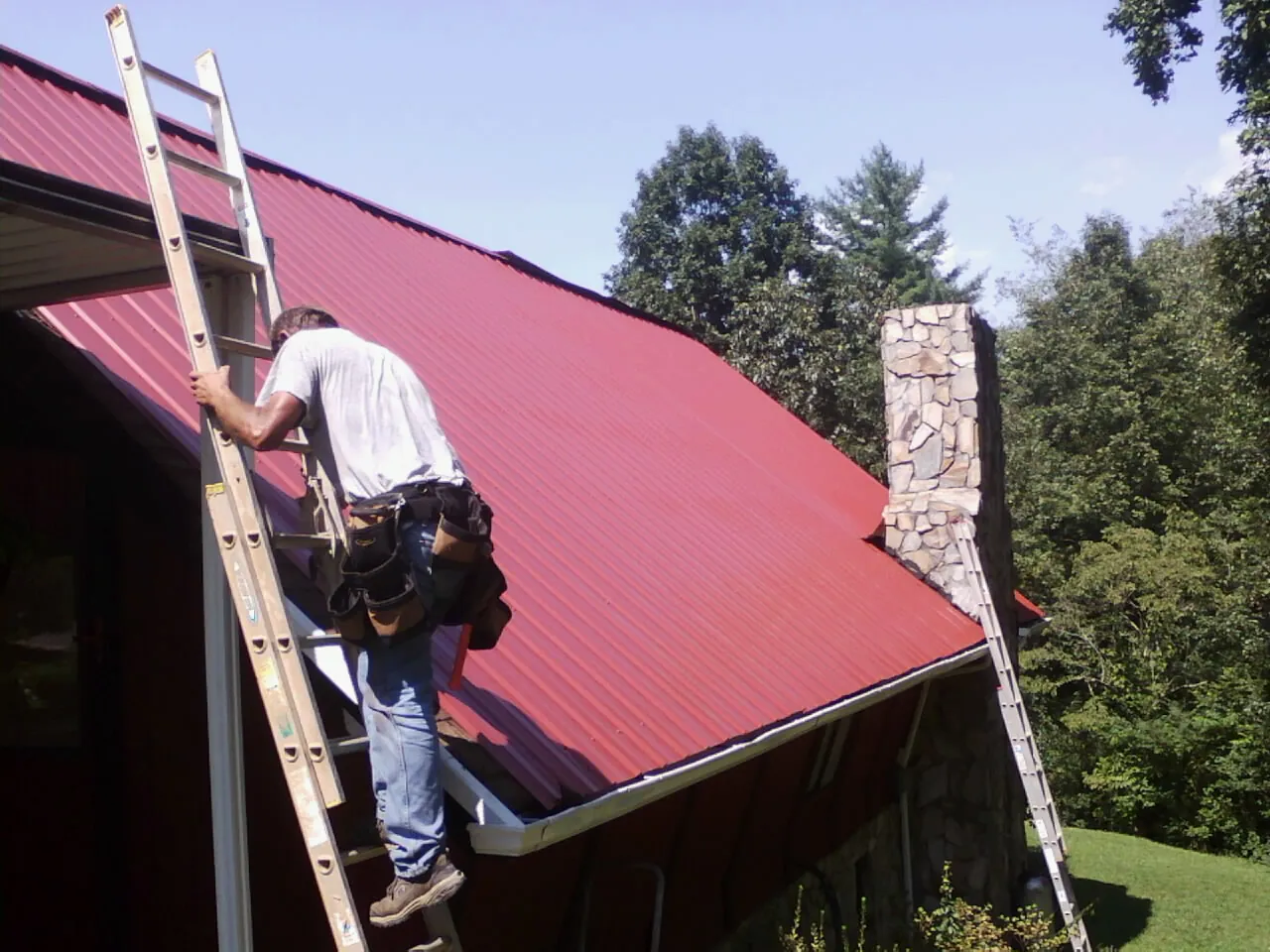 Roofer on a ladder beside a red metal roof and stone chimney — Soffit & Fascia Repair by Roofing Brothers in South Bound Brook